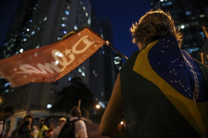 A person wrapped in a Brazilian flag stands beneath a banner in support of President Dilma Rousseff during a march in support of the president.