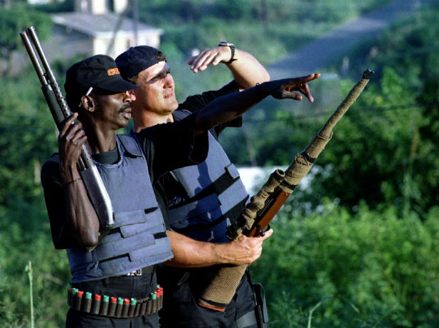 A South African mercenary (R) carrying a sniper rifle wrapped in camouflage netting, surveys a construction compound.