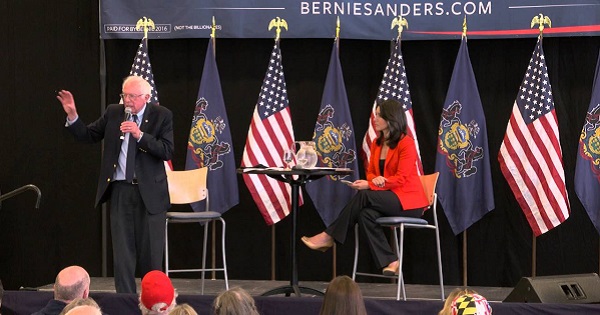 Bernie Sanders speaks at a rally while Tulsi Gabbard sits behind him.