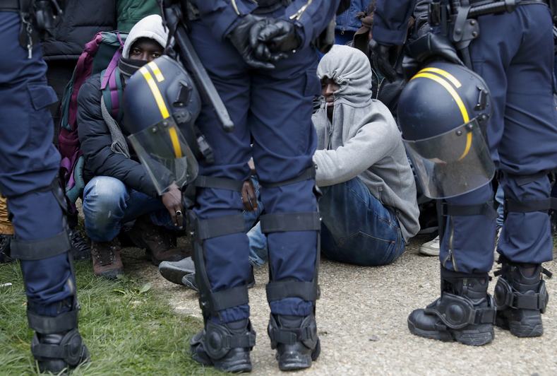 French police stand near as migrants with their belongings queue at the start of their evacuation and transfer to reception centers in France.
