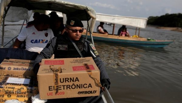 A police officer carries a package containing an electronic ballot box, in a community of 
